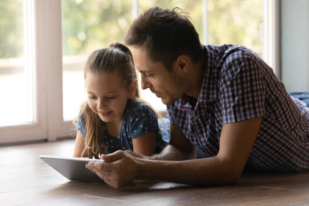 Shared hobby. Cute little daughter spend free time with young loving daddy lie on heated floor by window look at tablet screen. Junior schoolgirl read electronic book online browse web on pad with dadの写真素材