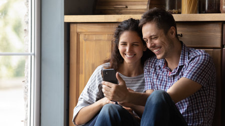 Happy millennial couple in love sit on warm floor at kitchen embrace looking on cellphone screen....