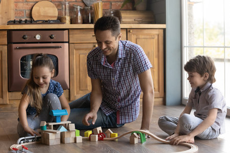 Offline activity. Smiling young daddy and diverse junior kids make build toy train station of colored wooden blocks. Male babysitter spend time with little boy girl play game at warm floor in kitchenの写真素材