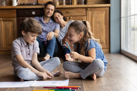 Preparing surprise. Friendly little kids brother sister sit on cozy heated floor whispering apart of mom dad. Cute children discuss plan to draw paint colorful picture card as gift for beloved parentsの写真素材