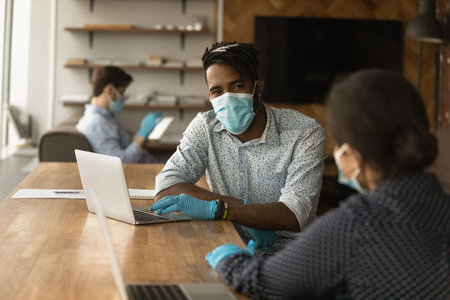 African American man in facial mask and rubber gloves from coronavirus talk with female colleague in office. Diverse employees in facemasks work together at coworking workplace. Covid-19 concept.の写真素材