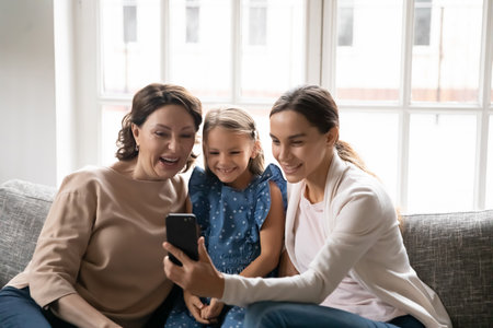 Overjoyed little girl with young mother and mature grandmother using phone, sitting on cozy couch at...