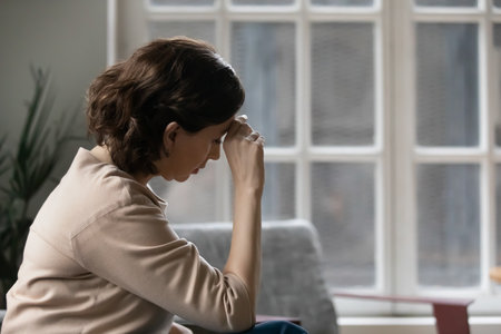 Rear view close up thoughtful mature woman touching forehead, suffering from headache, sitting on couch at home alone, pensive upset middle aged female lost in thoughts, thinking about problemsの写真素材