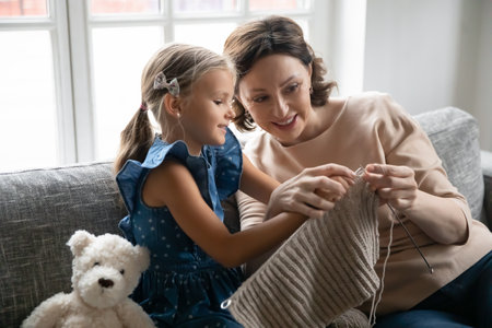 Close up smiling mature grandmother teaching little granddaughter knitting, sitting on cozy couch at home, positive happy middle aged woman and cute adorable girl engaged in creative activityの写真素材