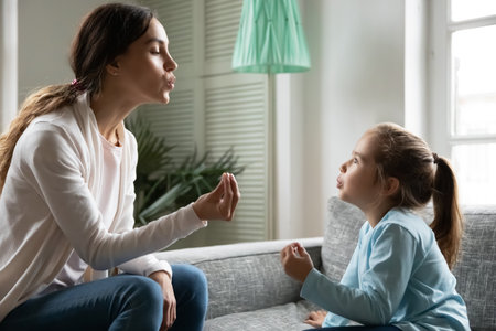 Close up little girl involved in speaking lesson with professional therapist, sitting on couch at home, caring mother teaching adorable daughter correct pronunciation, working with speech defectsの写真素材