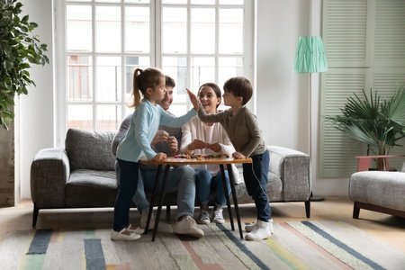 Happy parents with two kids having fun with colorful beads, sitting at table on cozy couch at home, cute adorable little daughter and son giving high five, family enjoying leisure time togetherの写真素材