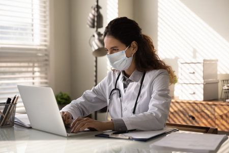 Young female doctor in white medical uniform and facial mask look at laptop screen consult patient online. Woman GP or physician in facemask have webcam consultation, work on computer in clinic.の写真素材