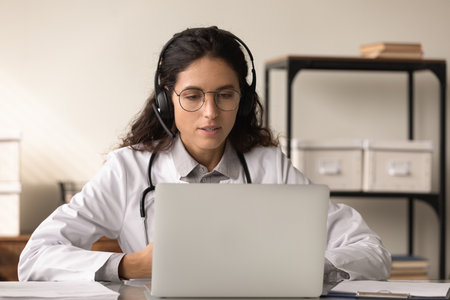 Focused young Caucasian female doctor in headphones look at computer screen talk on video call with patient. Woman GP in medical uniform and earphones have webcam online digital virtual with client.の写真素材