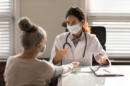 Female doctor in medical facemask measure blood pressure with electronic monitor in hospital. Caring woman GP help examine do checkup of elderly client in clinic. Healthcare, hypertension concept.の写真素材