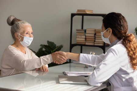Smiling female doctor and elderly patient in medical facemasks shake hands get acquainted at consultation in clinic. Woman GP and elderly client in facial masks handshake close health insurance deal.の写真素材