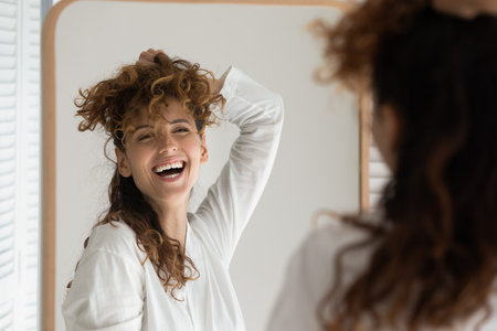 Overjoyed young Caucasian woman look in mirror at home bathroom have fun getting ready. Smiling millennial female try experiment with hairstyle or hairdo in bath. Hair beauty product concept.の写真素材