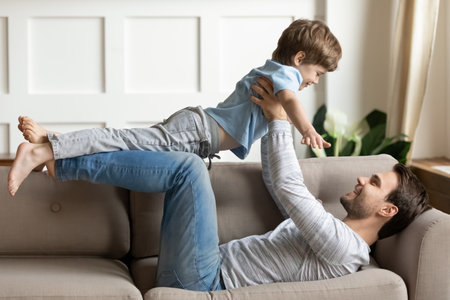 Loving young Caucasian father lying on couch at home play with excited little son on weekend. Happy caring dad imitate plane fly have fun feel playful engaged in game activity with small boy child.の写真素材