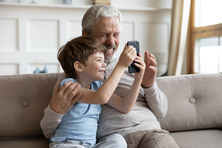 Smiling elderly grandfather and little 6s grandson have fun using modern smartphone relaxing at home together. Happy mature grandparent and small boy child talk on video call on cellphone.の写真素材