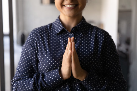 Crop close up of smiling woman hold keep hands in prayer feel superstitious religious at home. Happy millennial ethnic female pray to God, ask about good fate. Religion, faith, belief concept.の写真素材
