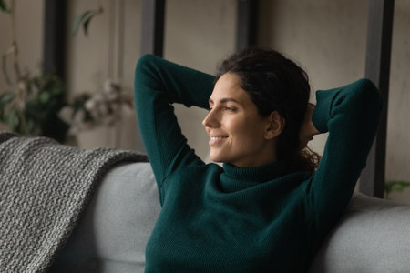 Smiling young Caucasian woman sit relax on couch at home look in distance dreaming or thinking. Happy female rest on sofa planning or visualizing, imagine good future or success. Vision concept.の写真素材