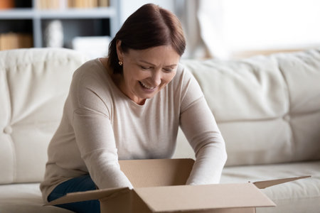 Close up smiling mature woman opening cardboard box at home, satisfied middle aged customer received awaited parcel with online store order, sitting on couch, good quick delivery service conceptの写真素材