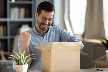 Happy 30s young man in eyewear looking inside of carton box, celebrating getting wished item or gift from internet store, having positive online shopping experience, fast postal delivery service.の写真素材