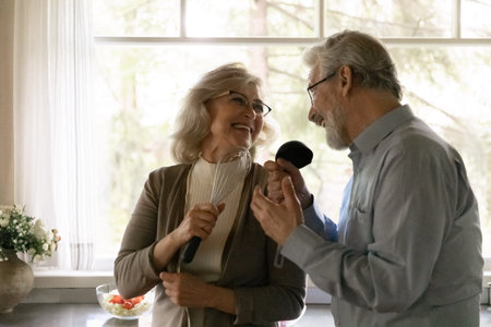 Overjoyed mature man and woman singing into kitchenware, holding beater and ladle, having fun in kitchen, enjoying leisure time, happy laughing elderly wife and husband dancing, listening to musicの写真素材