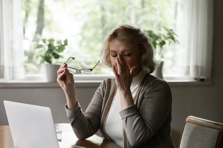 Exhausted stressed mature woman taking off glasses, touching massaging nose bridge, tired aged female suffering from eye strain after long laptop use, dry eyes syndrome, sitting at table at homeの写真素材