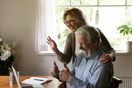 Overjoyed mature man and woman wearing glasses waving hands, looking at laptop screen, elderly couple wife and husband chatting online, making video call to relatives or friends at home togetherの写真素材