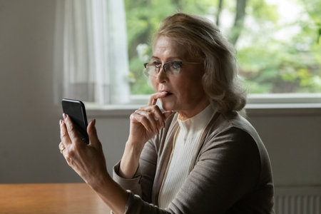 Close up thoughtful upset mature woman wearing glasses looking at phone screen, reading bad news in...