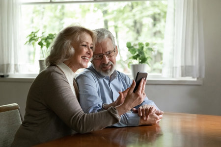 Happy mature man and woman using phone together, browsing apps, happy elderly wife showing funny video or news to husband, aged couple making video call, chatting or shopping online at homeの写真素材