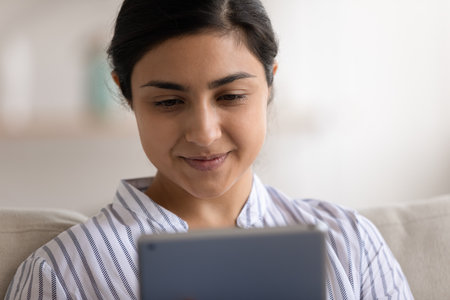 Using gadget. Close up shot of smiling indian female sit on couch watch good movie on tablet pc read blog enjoy video on device screen. Cropped view of positive young hindu lady electronic pad userの写真素材
