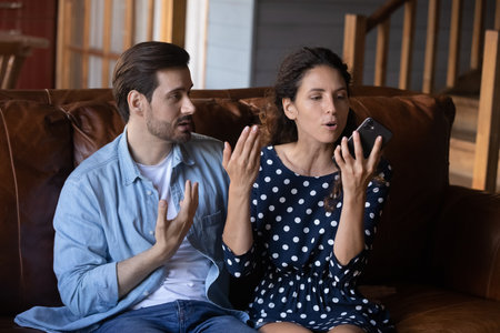Unhappy young Caucasian couple have family misunderstanding or fight over smartphone usage or addiction. Angry man and woman family or lovers quarrel using talking on cellphone gadget at home.の写真素材