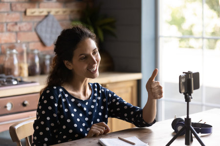 Smiling young Caucasian woman sit at table at home office record video blog on smartphone camera....
