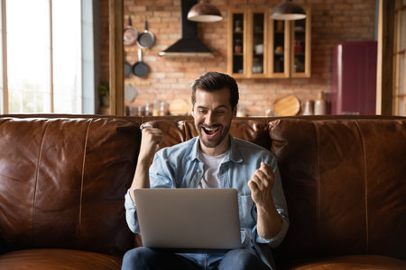 Overjoyed millennial Caucasian male relax on couch at home use laptop triumph win lottery online. Happy young man look at computer screen feel euphoric excited read good news or message on device.の写真素材