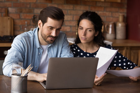 Young Caucasian couple look at laptop screen manage family financial paperwork paying bills online. Millennial man and woman work on computer, use web banking system for payment on gadget.の写真素材