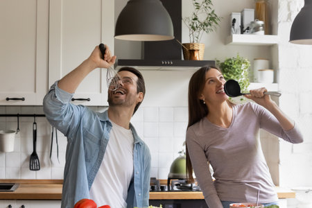 Funny cooking. Joyful family couple prepare food at home together having fun listening to music. Laughing young spouses playing pop stars dancing singing aloud in microphones made of kitchen utensilsの写真素材