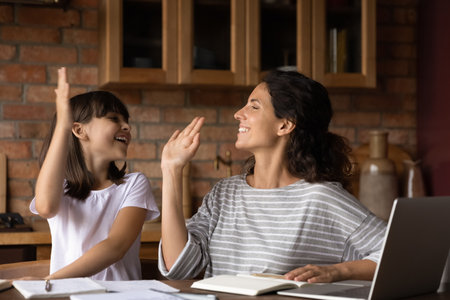 Happy mother and little daughter giving high five, studying online, doing assignments together, sitting at table with books and laptop at home, smiling teacher supporting schoolgirl pupilの写真素材