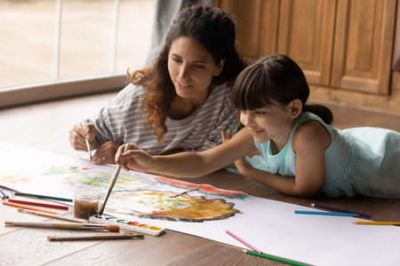 Close up smiling mum and little daughter painting colorful watercolors, lying relaxing on warm...