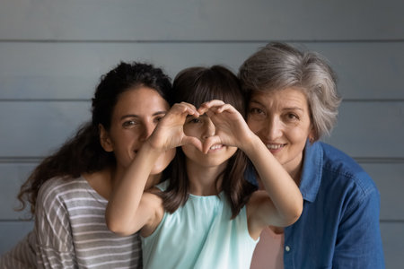 Head shot portrait little girl with young mother and mature grandmother showing heart gesture, looking at camera, three generations of women posing for family photo on grey studio backgroundの写真素材