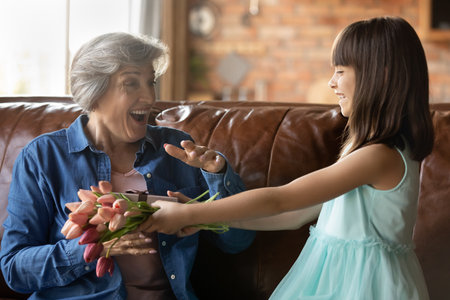 Close up little granddaughter congratulating mature grandmother, cute girl presenting flowers and gift box to overjoyed surprised older woman, family celebrating birthday or 8 march at homeの写真素材