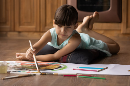 Close up little girl kid painting colorful watercolors on paper, holding brush, lying on warm wooden floor with underfloor heating, adorable child enjoying creative activity, creating pictureの写真素材