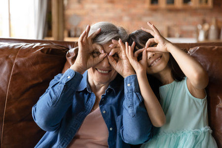 Head shot portrait of smiling mature grandmother and granddaughter making funny faces, binoculars glasses shape gesture, excited older woman with little girl looking at camera, having fun togetherの写真素材
