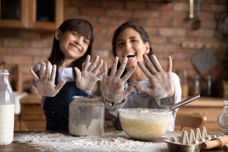 Close up happy young mother and little daughter showing hands in white flour at camera, cooking dough, smiling adorable girl with mum having fun in kitchen at home, preparing homemade pastryの写真素材
