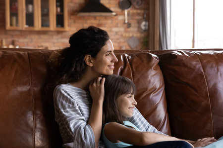Close up dreamy mother with little daughter hugging, looking to aside, dreaming, sitting on couch at home, smiling mum and adorable girl child relaxing, visualizing future, enjoying weekendの写真素材