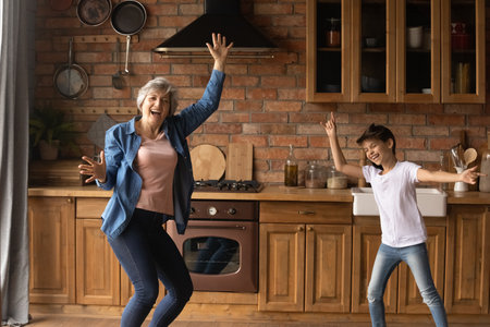Overjoyed mature woman with little girl dancing in kitchen at home, having fun, aged grandmother and adorable granddaughter moving to music, listening to favorite song, involved in funny activityの写真素材
