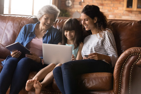 Happy three generations of women having fun with gadgets together, browsing apps, sitting on cozy couch at home, smiling little girl with mother using laptop, mature grandmother holding tabletの写真素材