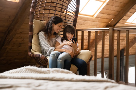 Happy mother with little daughter using phone, relaxing in cozy rattan armchair at home together, smiling mum and adorable girl looking at phone screen, surfing internet, chatting or shopping onlineの写真素材