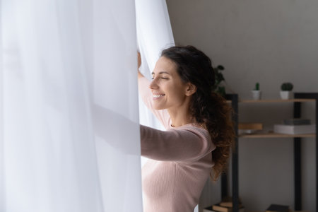Side view happy young beautiful caucasian woman opening curtains, enjoying morning cityscape view, welcoming new sunny day, feeling motivated inspired alone at home or hotel, dreaming indoors.の写真素材