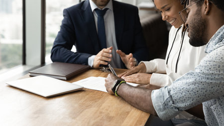 Confirming deal. Close up of happy african husband wife signing up contract in presence of lawyer financial advisor. Young black couple buy real estate make investment after getting agent consultationの写真素材