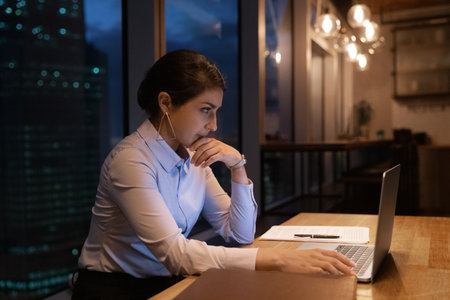 Thoughtful indian businesswoman work by laptop at studio apartment or modern office while night cover city outside panoramic window. Pensive young hindu female read document online on computer screenの写真素材