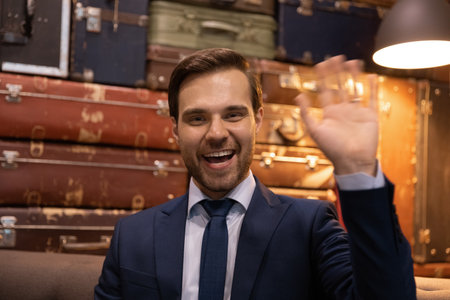 Headshot portrait of young smiling bearded businessman in formal suit look at camera wave hand. Millennial male manager consultant call client from modern office timeclub greet customer start talkの写真素材