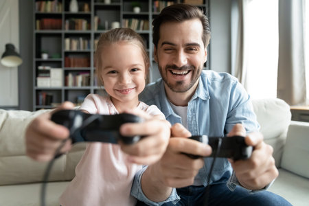 Happy small 7s kid girl playing online video games with young father, sitting together on sofa....