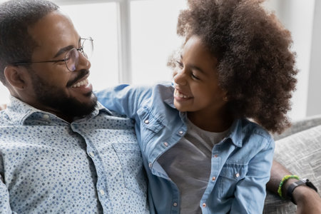 Close up overjoyed African American father wearing glasses and daughter having fun, hugging, relaxing on cozy couch at home, happy dad with adorable child girl enjoying leisure time togetherの写真素材