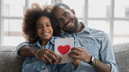 Head shot portrait smiling African American father hugging daughter, holding handmade greeting card with heart, looking at camera, loving little girl congratulating dad with birthday or fathers dayの写真素材